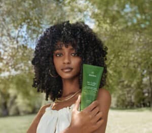 A woman with curly hair holding a bottle of aveda be curly product in a sunny park.