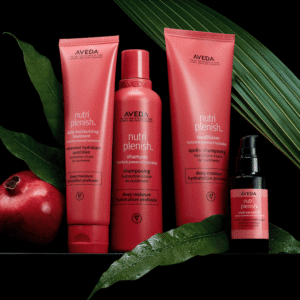 A set of Aveda Nutriplenish hair care products, including shampoo, conditioner, daily moisturizing treatment, and a small bottle, arranged with foliage and a pomegranate on a black background.