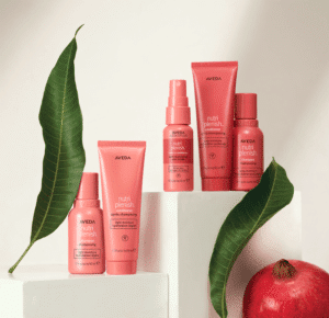 A set of five Aveda Nutri Plenish hair care products arranged on white blocks with a pomegranate and large green leaves. The products are in pink packaging and include shampoo, conditioner, and spray.