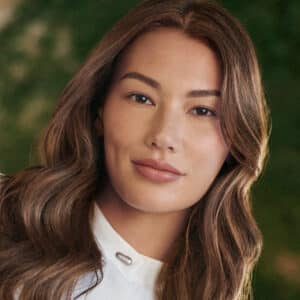 A woman with long wavy brown hair and a white blouse smiles softly, standing against a green, natural background.