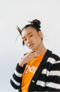 Person wearing an orange shirt and black-and-white striped sweater, with orange eye makeup and hair styled in two buns, posing against a plain background.