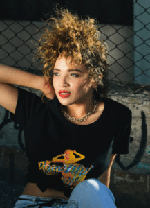 Person with curly hair, wearing a black T-shirt with colorful print, sits outdoors against a chain-link fence.