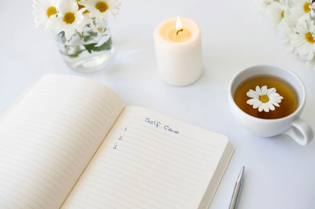 Open notebook with "Self Care" list, lit candle, pen, daisy flowers in a vase, and cup of tea with a daisy floating on top.