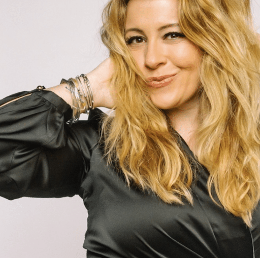 Woman with wavy blonde hair wearing a black blouse and silver bracelets, posing with one hand behind her head against a plain background.