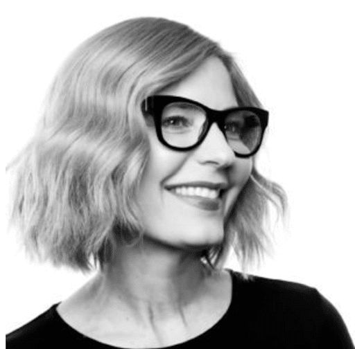 A woman with wavy bob hairstyle and large glasses smiles, looking to the side. She is wearing a dark top. The image is in black and white.