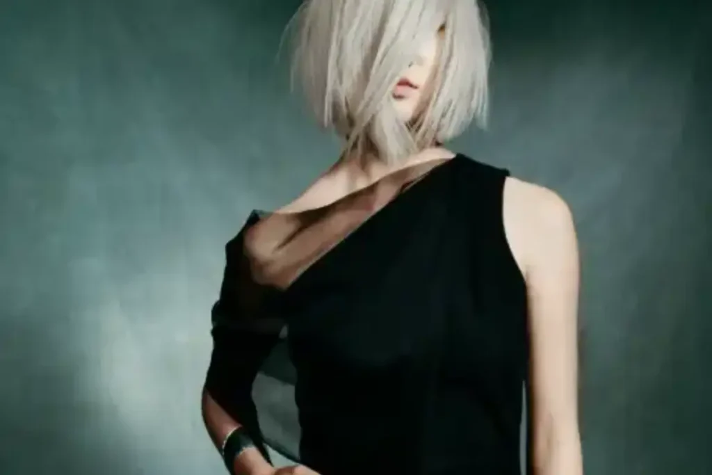 Woman with platinum blonde bob hairstyle wearing an asymmetrical black dress stands against a dark, blurred background, embodying self-care and elegance reminiscent of an Aveda Botanical Repair transformation.