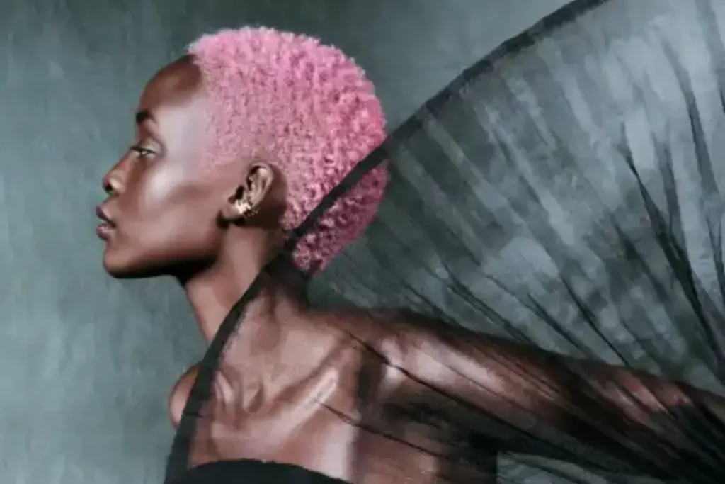 A person with short pink hair poses in profile against a dark background, draped in a sheer black fabric—capturing a quiet moment of self-care.