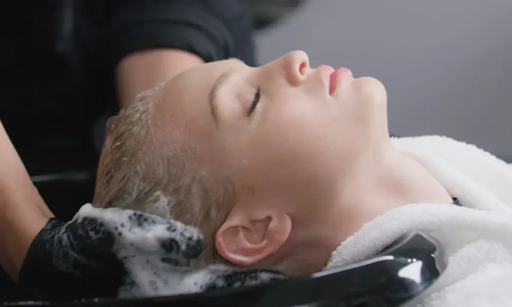 A person with closed eyes reclines at a salon sink while their hair is washed with shampoo, creating lather.