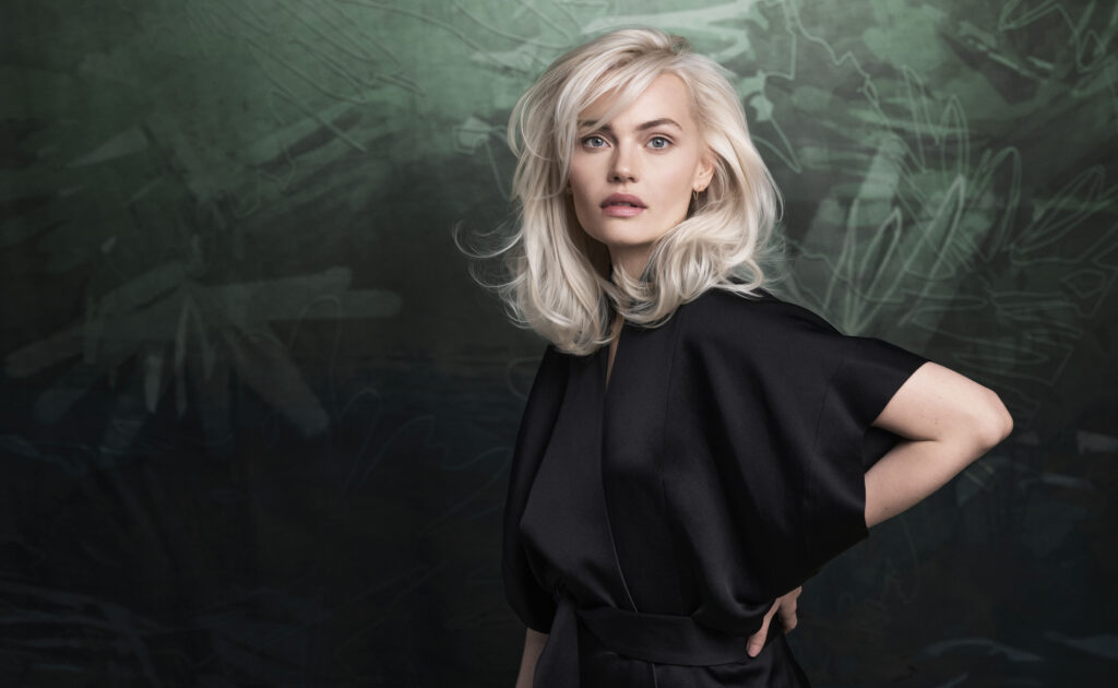 A woman with platinum blonde hair wearing a black outfit stands against a dark green and black abstract background, gazing at the camera.