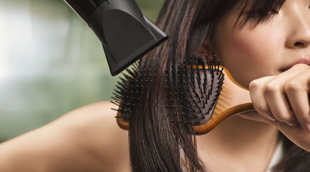 A person blow-drying their straight, dark hair with a hair dryer while brushing it with a wooden paddle brush.