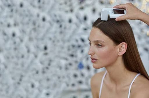 A person uses a handheld device to scan the scalp of a seated woman with long brown hair, highlighting the benefits of scalp exfoliation, against a blurred patterned background.